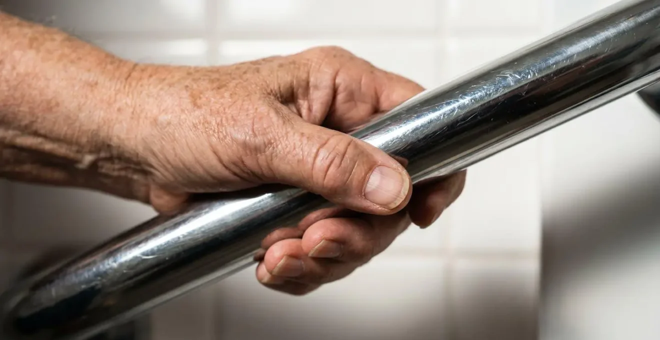 Gros plan sur une main senior posée sur une barre d'appui chromée dans une salle de bain épurée